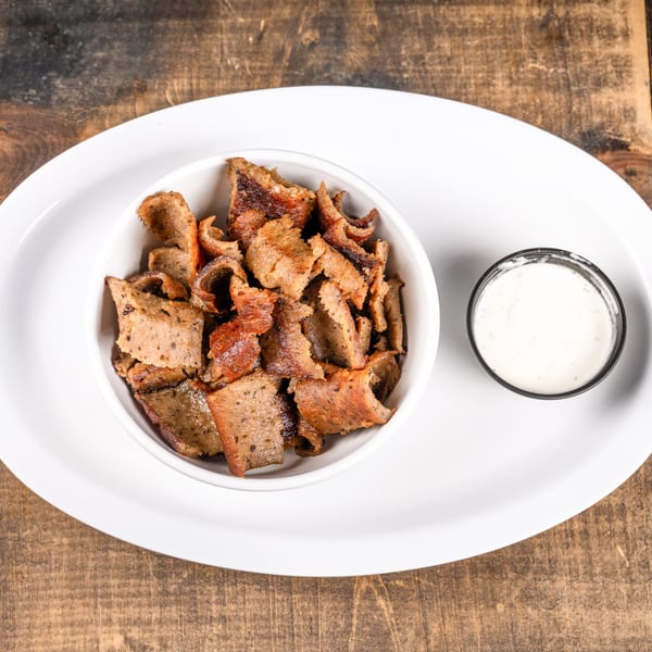 A white bowl filled with gyro meat and a small cup of tzatziki sauce on a white plate.