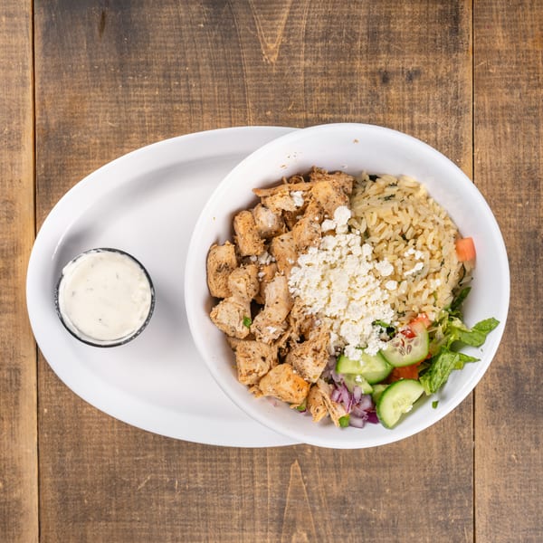Chicken and rice bowl with feta, salad, and a side of dressing on a wooden table.