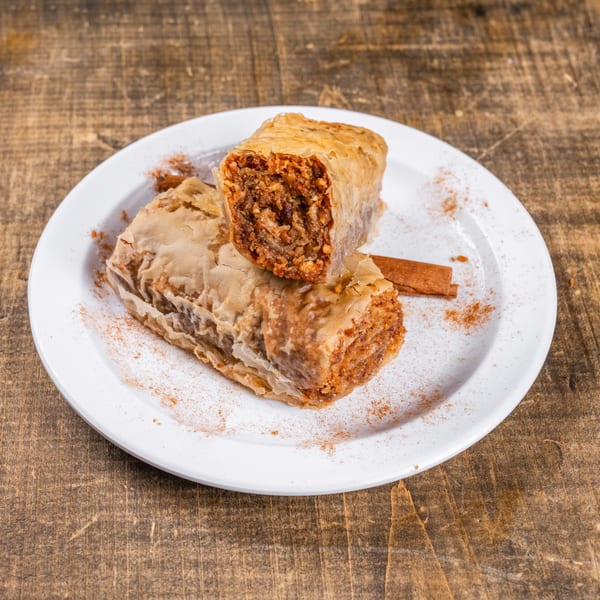 Two pieces of baklava, one cut open, on a white plate with cinnamon sticks and powder.