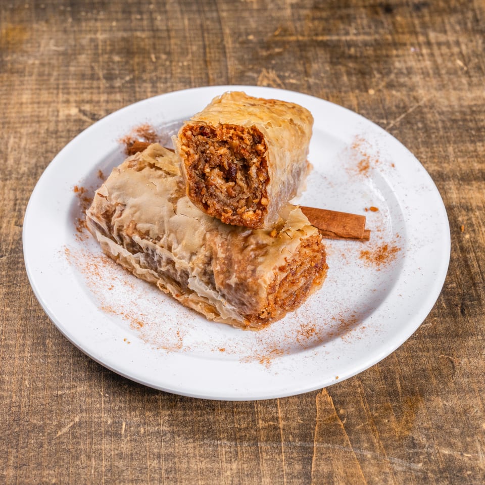 Two pieces of baklava, one cut open, on a white plate with cinnamon sticks and powder.